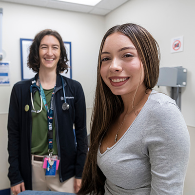 In the foreground, a teen girl smiles directly at the camera. She has long brown hair with blonde highlights and is wearing a gray shirt and gold necklace. Slightly out-of-focus in the background is a tall female doctor with a stethoscope and ID badge around her neck. She is smiling as well.