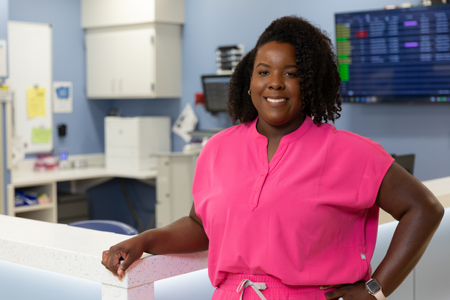 Register nurse chelsea krivanek in hot pink scrub top and pants posing against nursing station counter