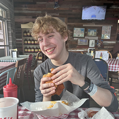 A teenage boy sits at a restaurant table holding a chicken sandwich, with a food basket and drink on the table.