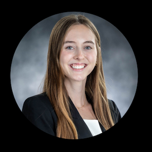 Professional headshot of a person with long hair wearing a dark blazer and light-colored top, photographed against a gray studio background.