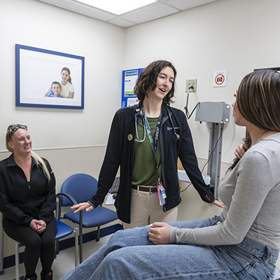 A tall, female doctor with chin-length brown hair stands facing a patient. The patient is a teenage girl with long dark hair with blonde highlights. She is wearing a gray shirt and jeans and sitting on an exam table. The patient’s mom sits off to the side, smiling and watching the interaction.