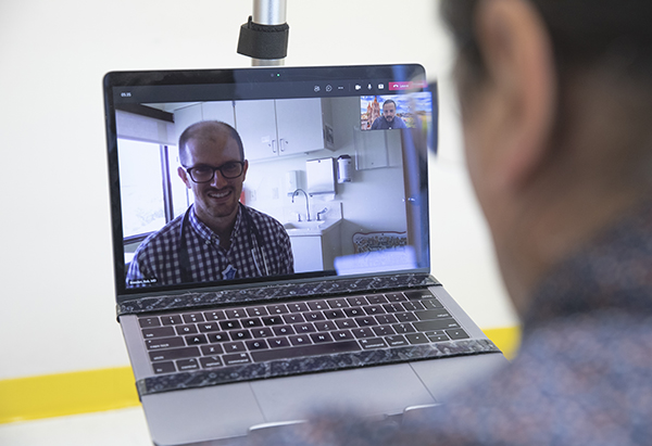 a CM physician’s face is on a laptop computer screen smiling and a Cultural Language Coaching Program male staff member is also on the laptop computer screen in a small box in the top right-hand corner. Both are virtually meeting with a man who is holding the laptop computer, with only the back of his head shown.