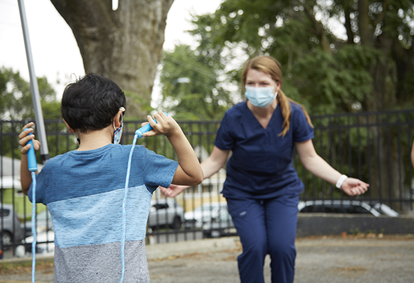 a female dressed in clinical navy-blue scrubs with mid-length sandy brown hair is holding her hands outstretched pretending to hold an invisible jump rope, while a little boy holds a blue jump rope in his hands, with the jump rope facing his back.