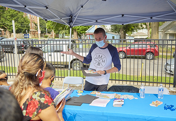 a male in a mostly white t-shirt w/navy blue mid-length sleeves with a CM logo on the front, is holding a black folder and colorful kiddie stickers in one hand. There are 5 people lined up at his table where he is handing out pamphlets and information on healthcare