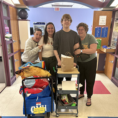 A teenage boy stands in a hospital hallway with three adults beside two rolling carts filled with personal items and supplies.