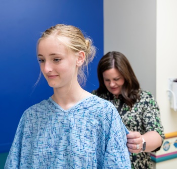 A teenage girl with blond hair wearing a hospital gown has her back measured by a clinician.