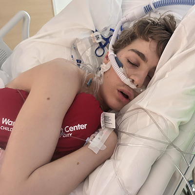 A teenage boy lies in a hospital bed with medical tubing visible and holds a heart shaped pillow against his chest.