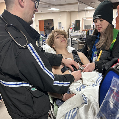 A teenage boy lies on a hospital stretcher while clinicians adjust monitoring equipment in a clinical care area.