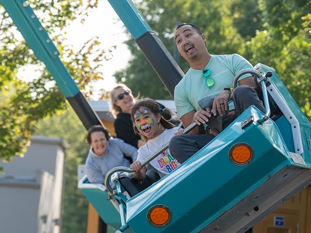 Child riding a teal amusement park ride with three accompanying riders seated behind, outdoors among trees on a sunny day.