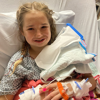 A young teen with blond braids is propped up in a hospital bed. She is wearing a hospital gown and has IVs in both hands and a large ice pack near her face. She is smiling but also experiencing some swelling from surgery on her jaw.