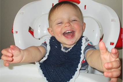 Dallin Mecham as a toddler sitting in a high chair, wearing a bib and smiling with his arms outstretched.