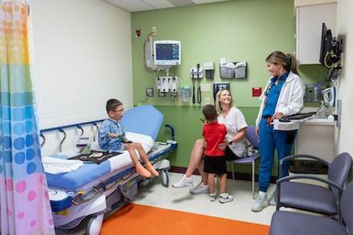 male child patient sitting on hospital bed in blue hospital gown. Patient mother in white and sibling in red shirt all facing provider in blue scrubs and white coat