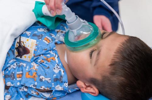 male child patient laying down in blue hospital gown. Child's eyes are closed and a clinic staff member is holding and anesthesia mask to child's face.