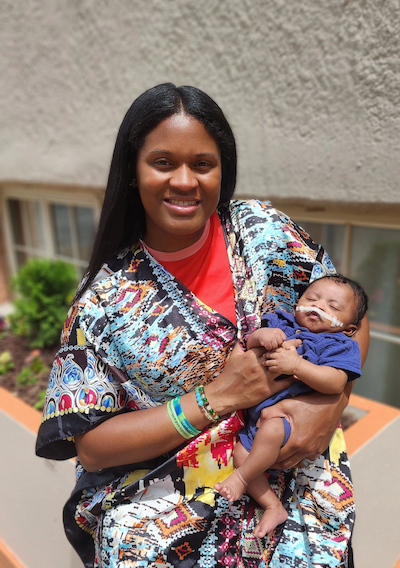 Aisha holds baby Wisdom. They are outdoors.