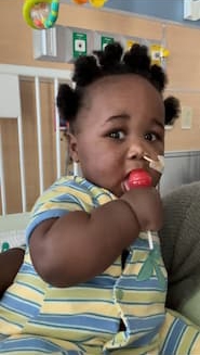 A baby in striped onesie eats a sucker. He has a tube coming from his nose, and hospital equipment is in the background.
