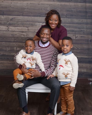 A family (a mother, father and two little boys) poses for a formal portrait. The father is sitting in a chair, holding the youngest boy. To his right, the older boy is standing with his hand on his father’s knee. The mother is standing behind the chair with her hands folded on top of it. They are all smiling, and the boys are wearing matching outfits: Christmas sweaters with Santa and reindeer decorations over denim shirts and orange pants.