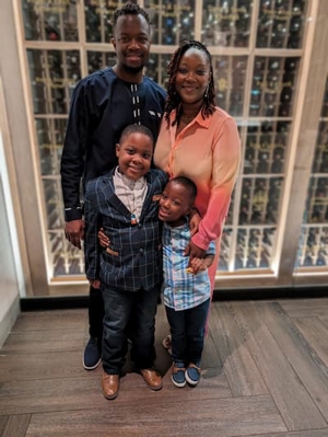 A family stands together, smiling at the camera. They are dressed up for a celebration. The father stands back left with the mother at his side. In front, the boy on the left is wearing a suit coat and has his arm around his little brother, who is shorter than him and wearing a striped shirt.