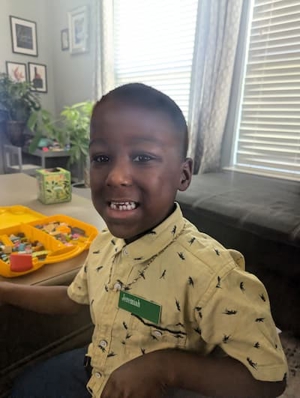 A young boy with a nametag reading “Jeremiah” opens his mouth to show off his missing lower tooth. He is wearing a yellow button-up shirt with tiny T-rexes printed on it, and there are toys on the table in front of him.